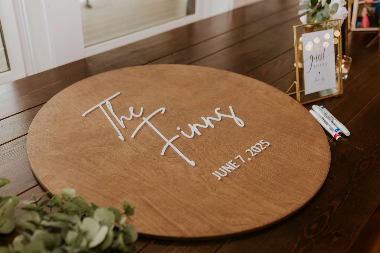 Round wooden wedding guest book sign reading The Finnys June 7 2025 in white script calligraphy.