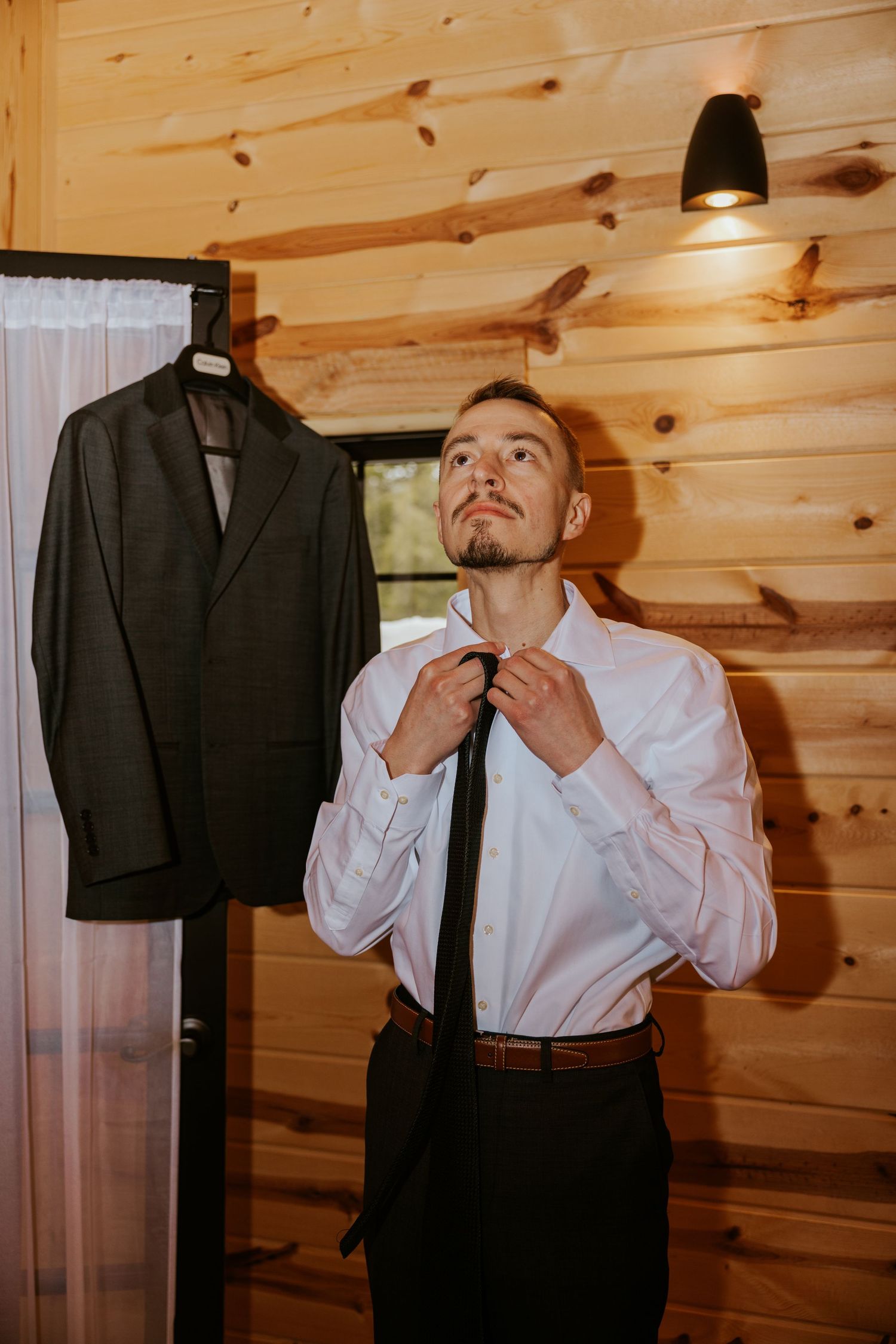A groom adjusts his tie while getting dressed in a wood-paneled room with a suit jacket hanging nearby.