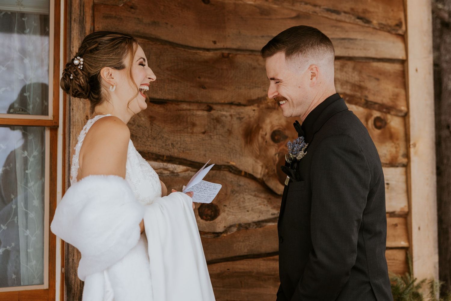 Bride reads wedding vows to groom outside rustic log cabin venue, both smiling during intimate ceremony.
