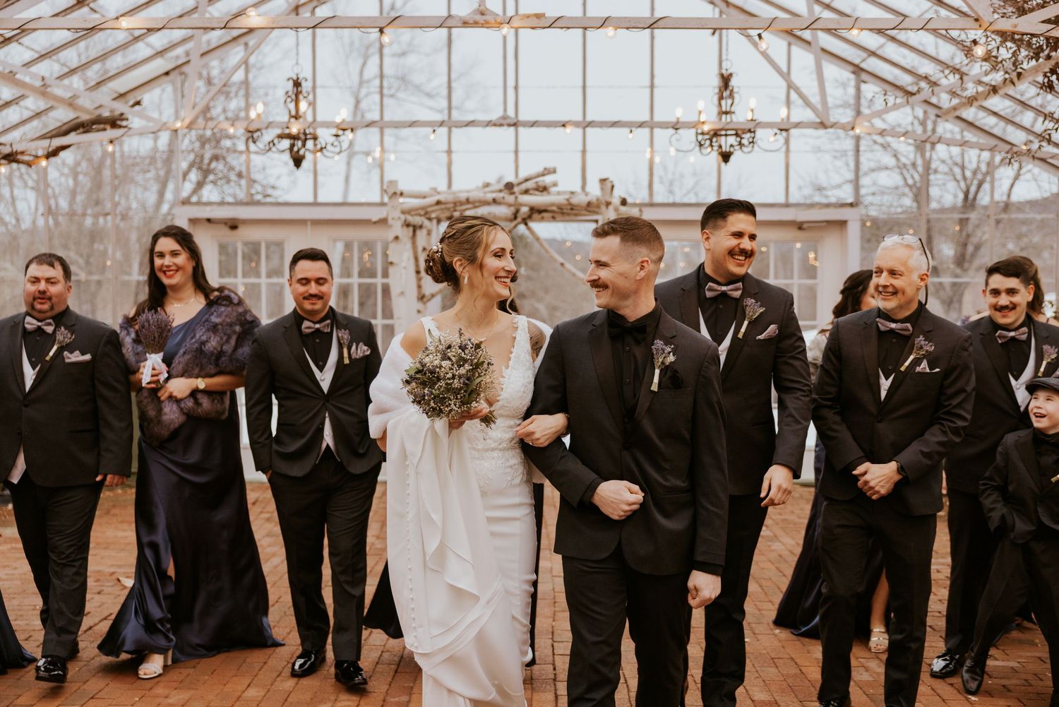 Wedding party in black attire laughs together inside a greenhouse venue with the bride holding a bouquet.