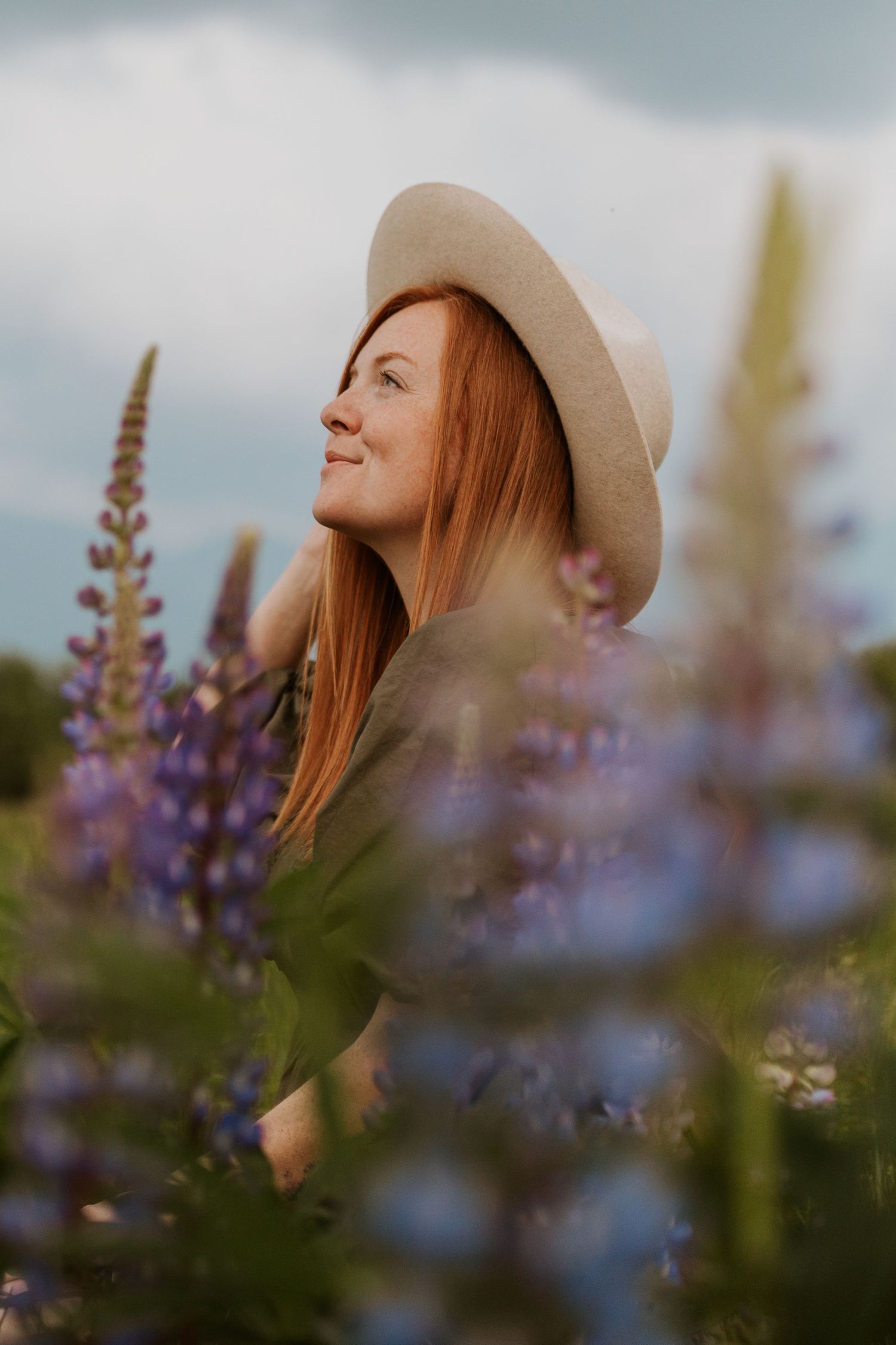 Smiling redhead woman in a beige hat surrounded by blooming purple lupine flowers under a cloudy sky.