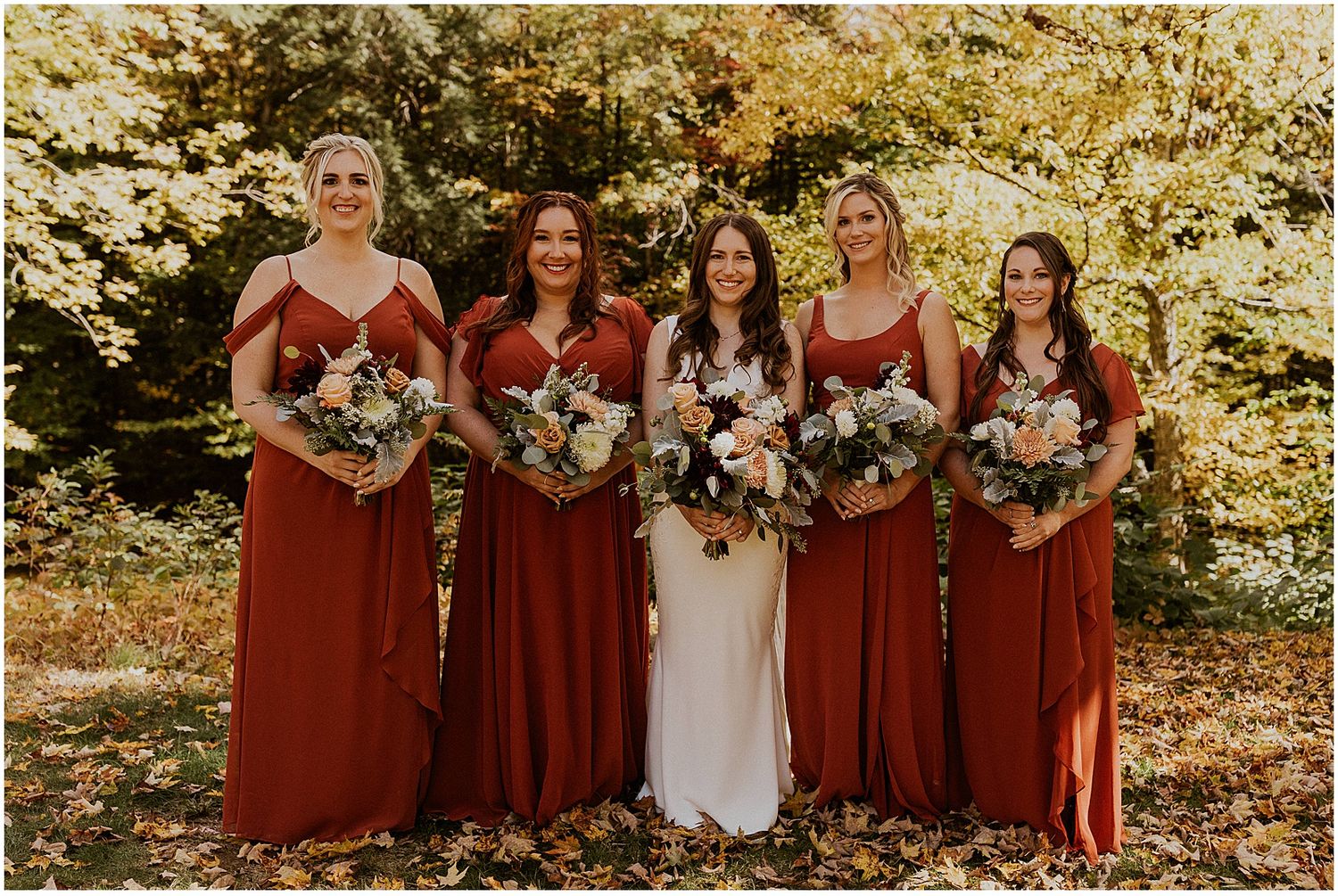 A bridal party wearing burgundy dresses poses together in autumn foliage.