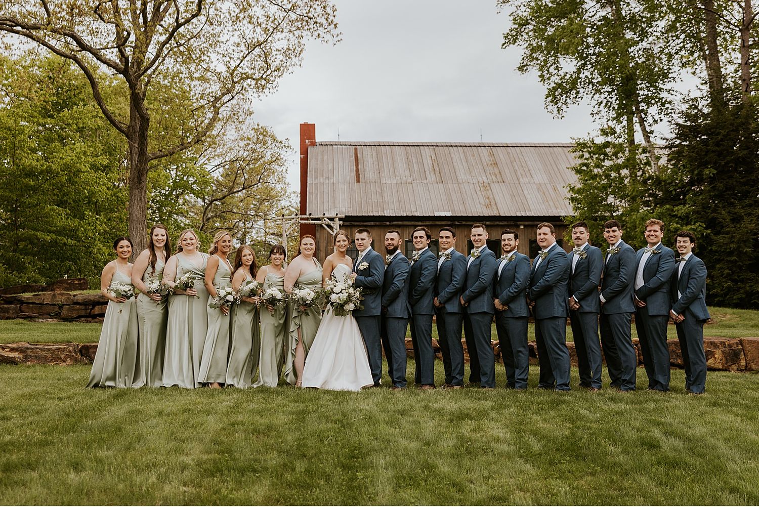 Large wedding party in navy and neutral colors posed outdoors on grassy lawn.