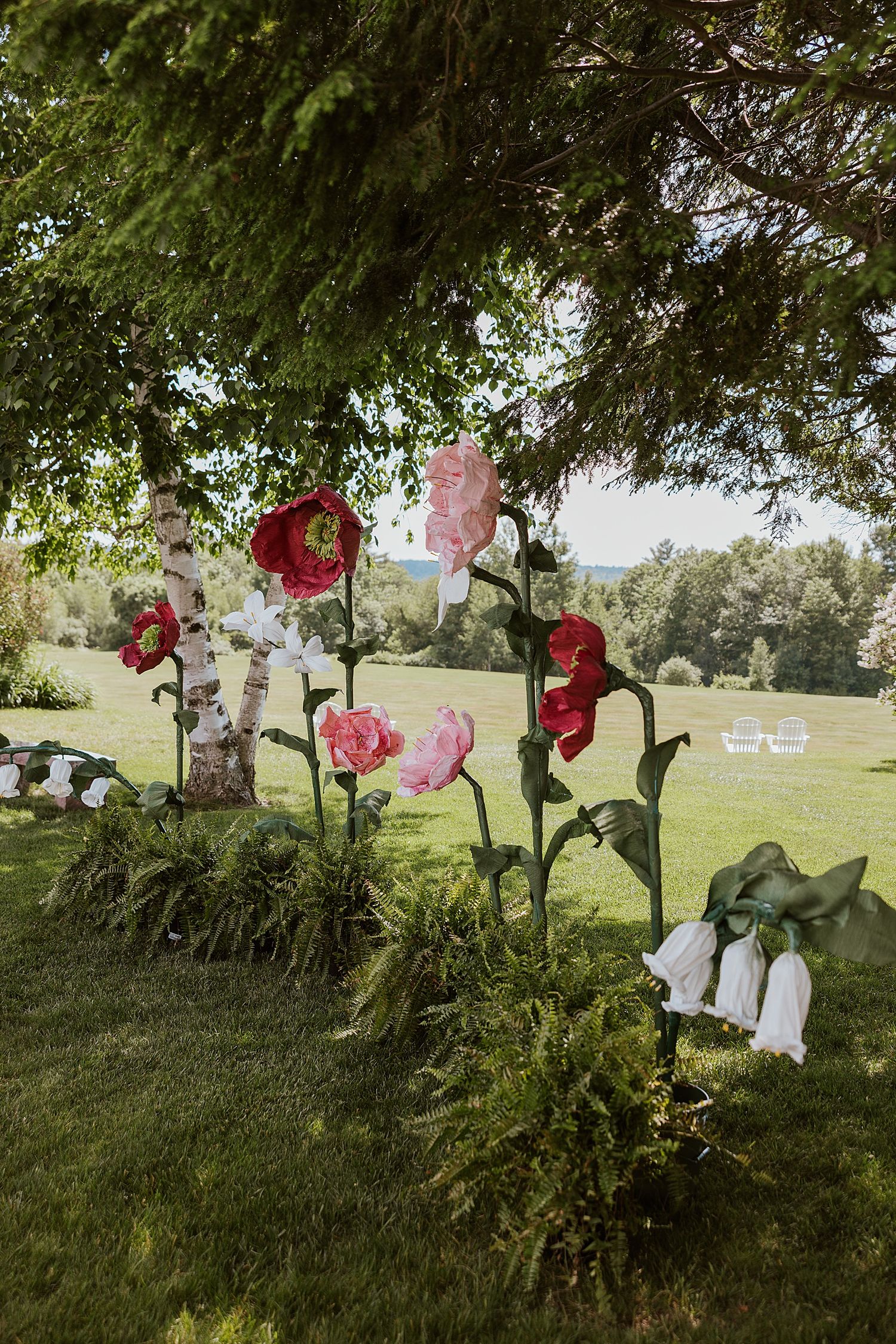 Large decorative paper flowers in pink and red displayed outdoors on green lawn under tree canopy at garden event.