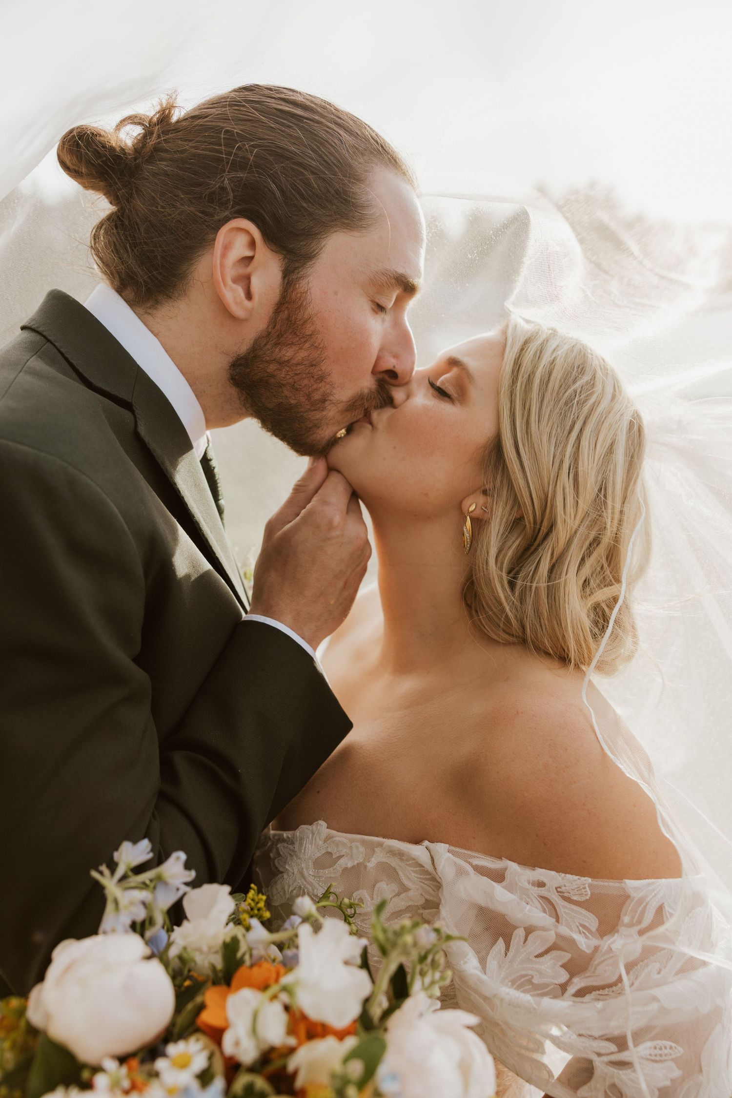 Bride and groom share romantic kiss under veil with colorful floral bouquet in foreground.