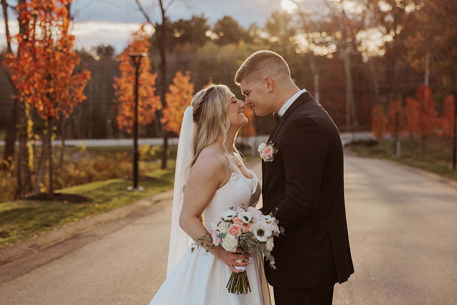 Bride and groom share a romantic kiss on a tree-lined road surrounded by vibrant autumn foliage at golden hour.