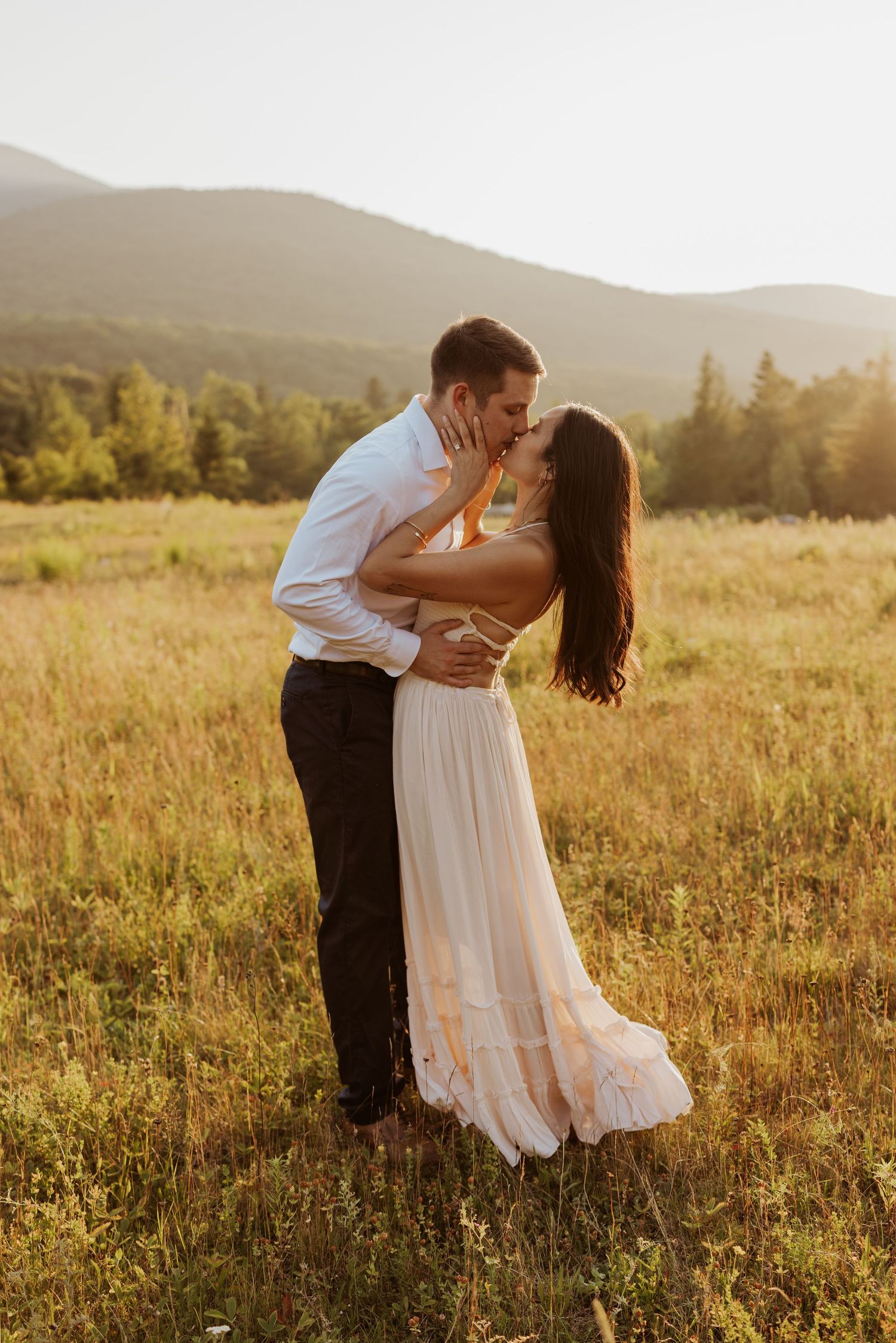 Couple shares romantic kiss in sunlit field with rolling mountains behind them.