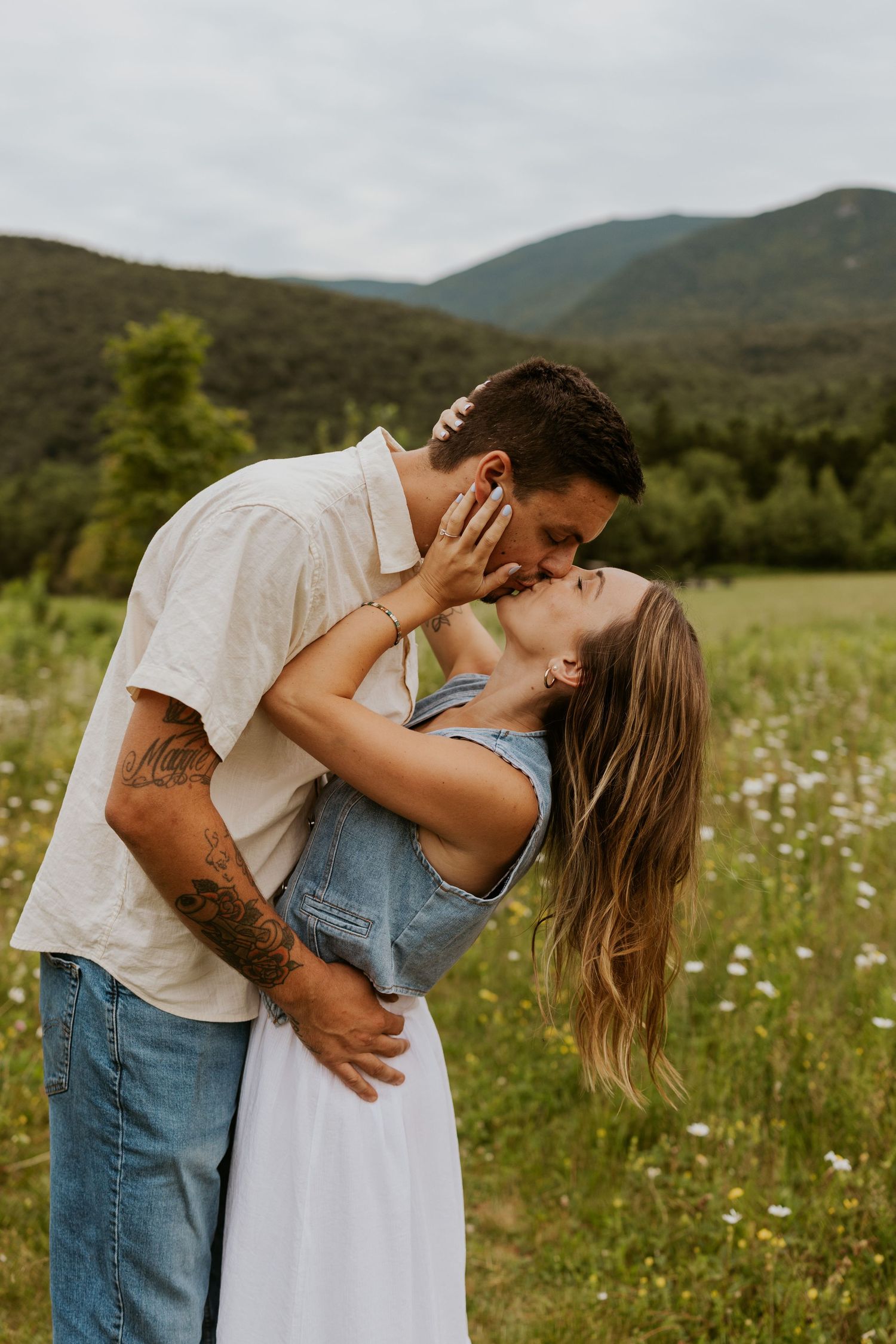 Passionate couple kissing in summer meadow, woman leaning back with mountains visible behind them.