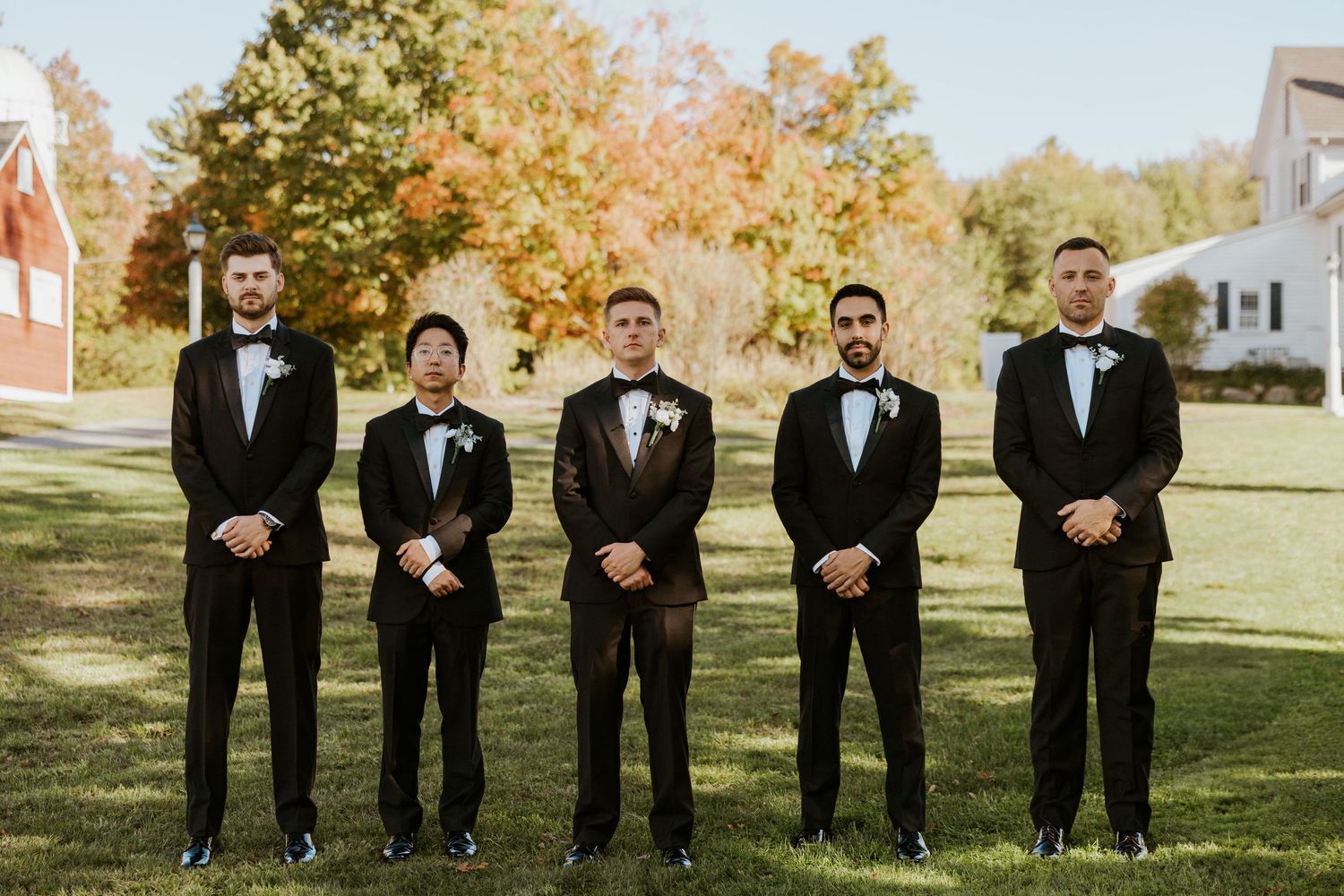 Groom and four groomsmen pose in matching black tuxedos on green lawn with autumn trees.