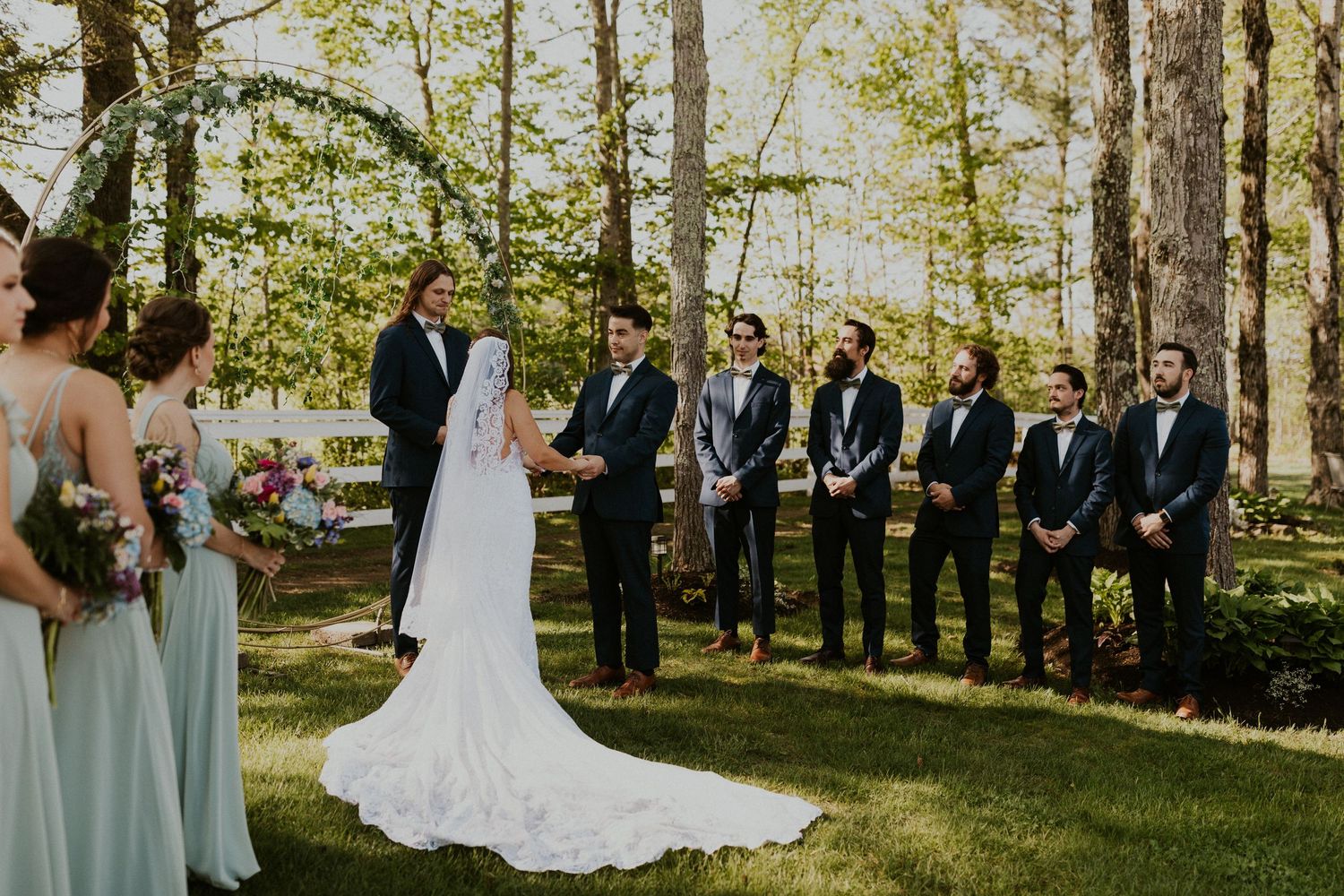 Chrissy and Mike holding hands as their wedding ceremony begins. It is a lovely spring day with the sun shining.