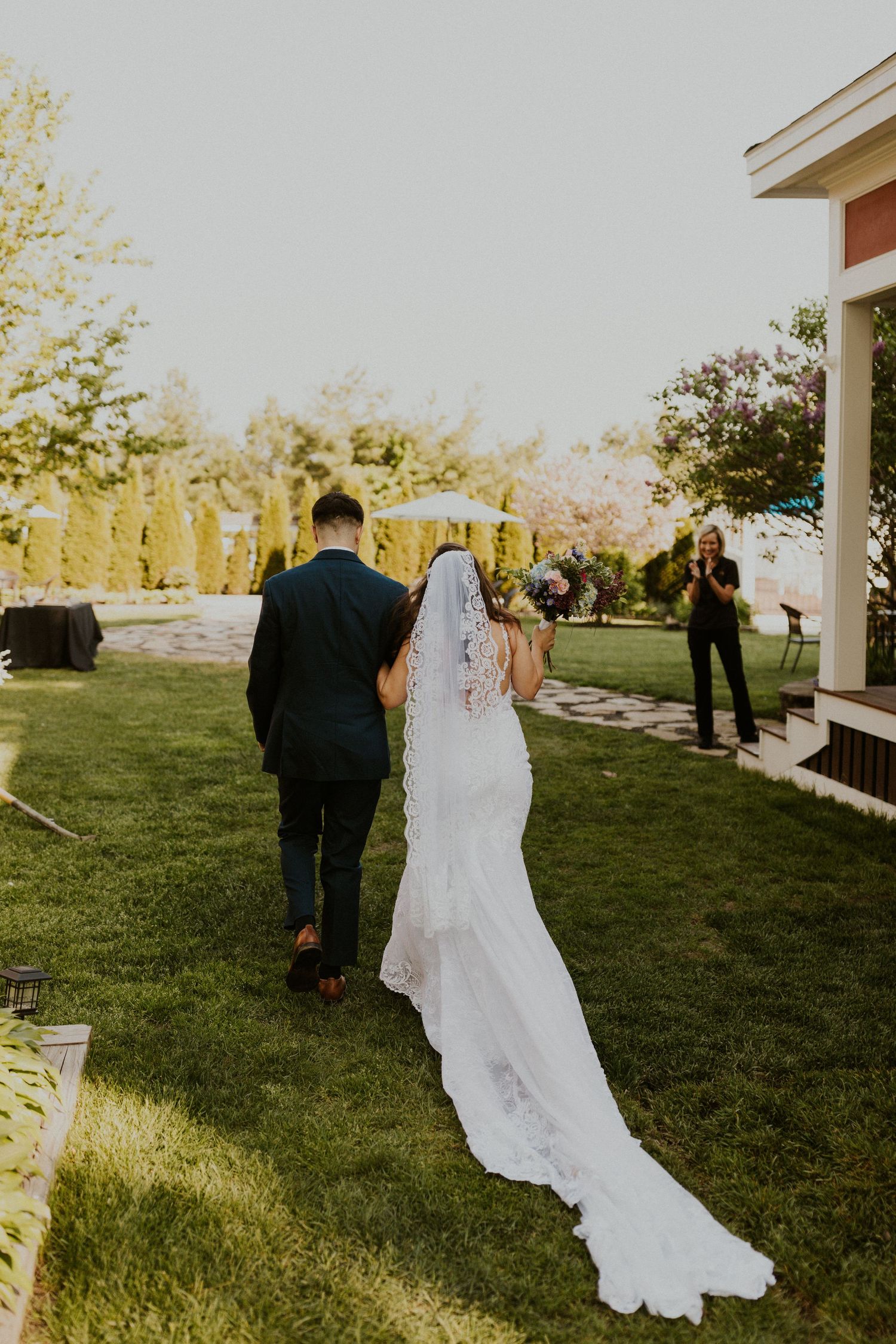 Wedding couple walks across a sunlit lawn with guests in the background.