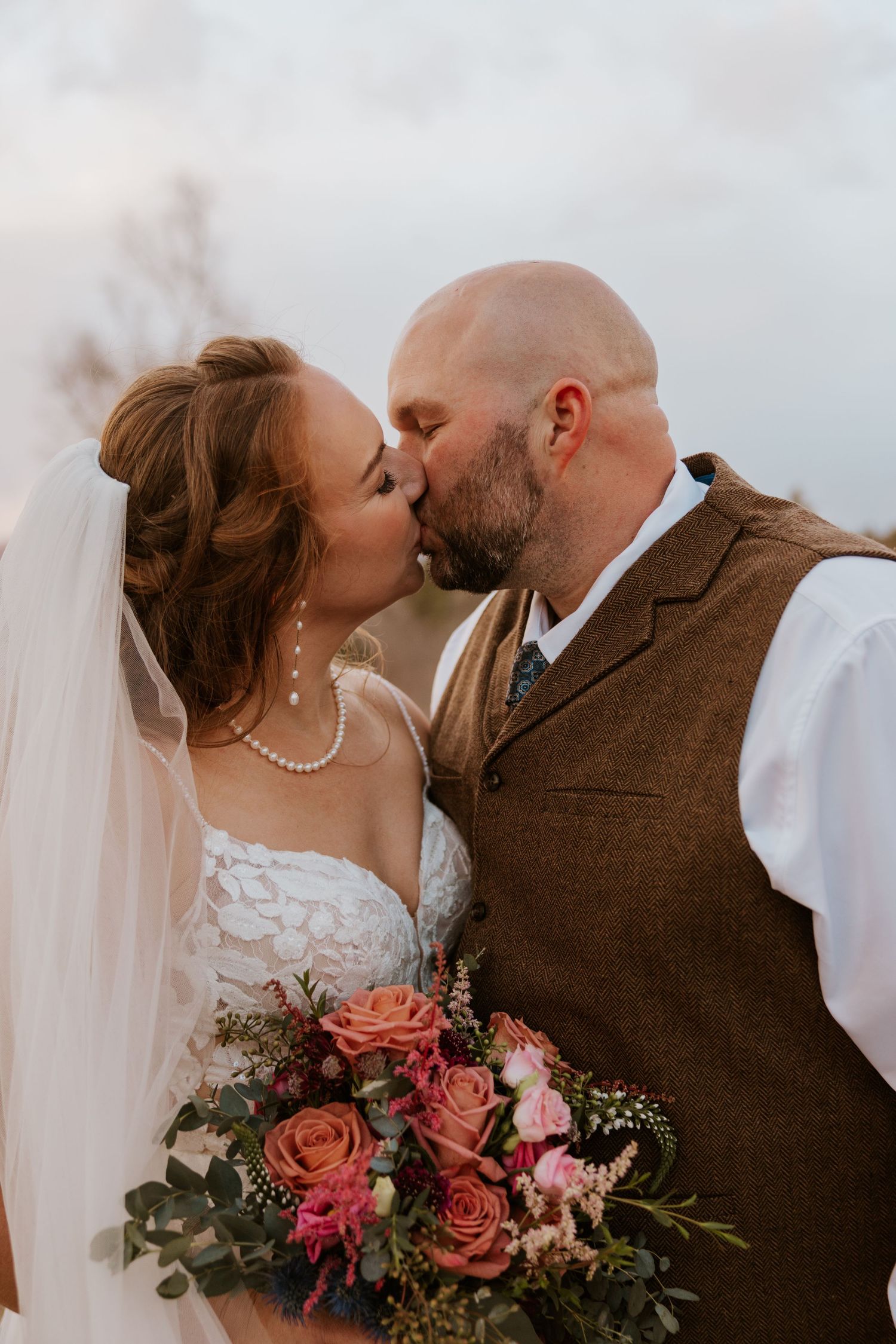 Romantic sunset portrait of newlyweds with coral roses and greenery bouquet.