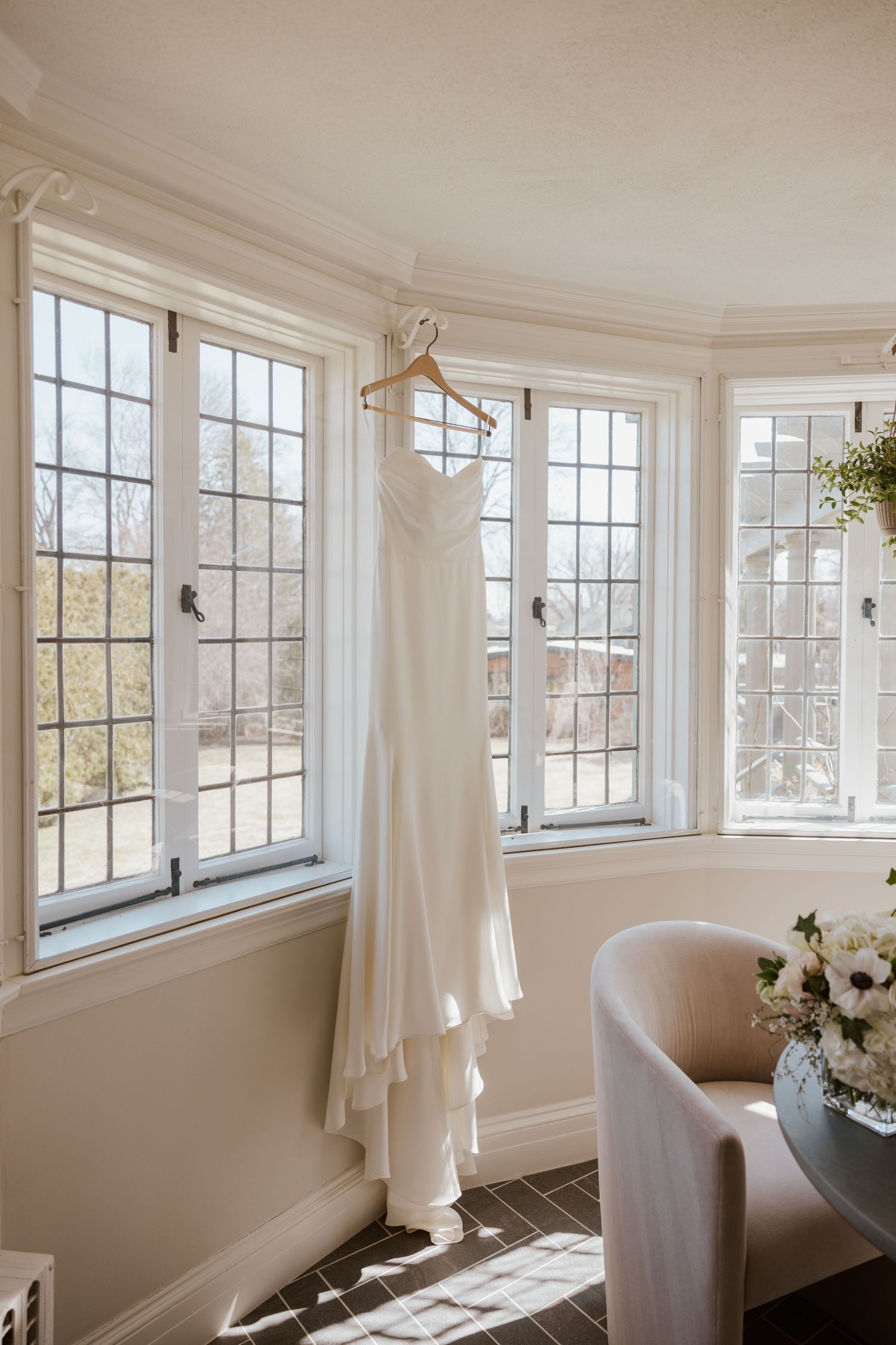 A white wedding dress hangs in a bright bay window with sunlight streaming in, next to a gray upholstered chair and flowers.
