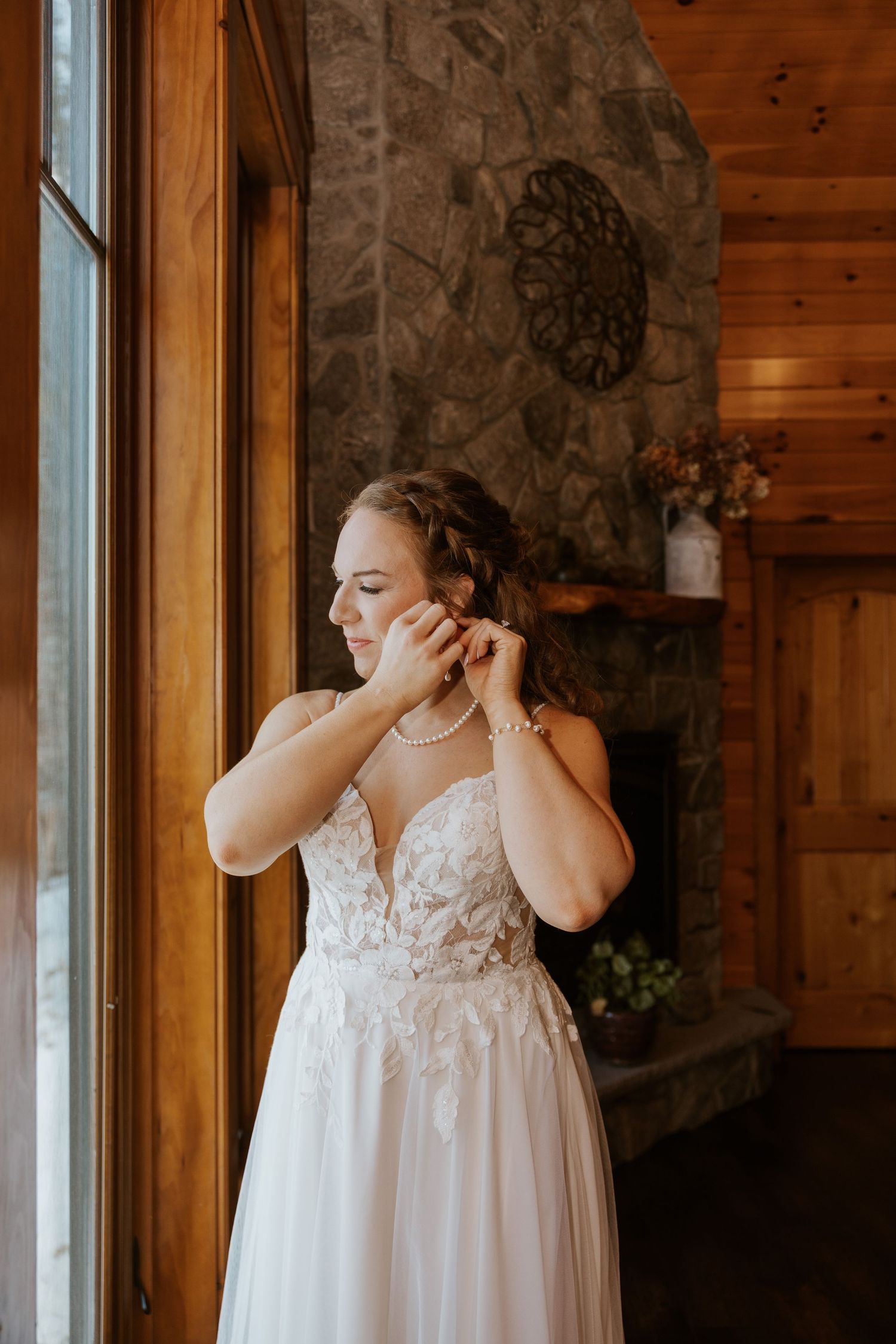 A bride in a white lace dress stands near windows in a rustic cabin-style room.