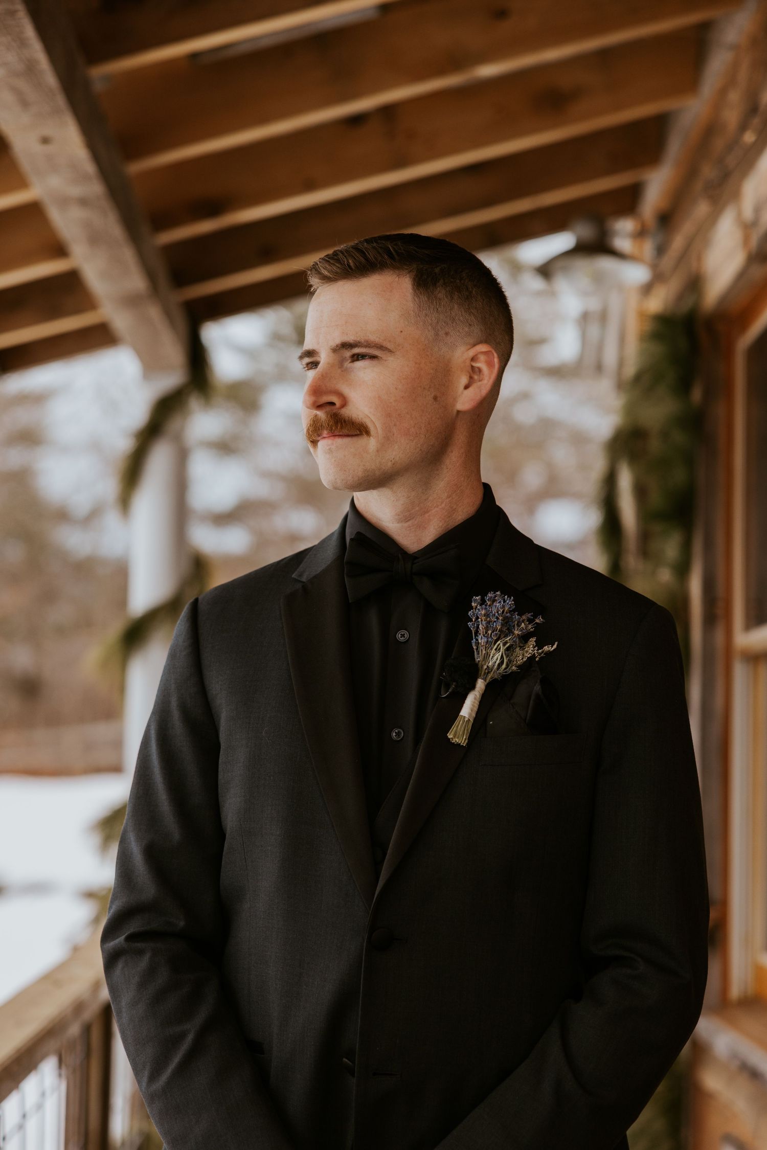 Dapper groom in black suit with boutonniere standing on snowy rustic cabin porch.