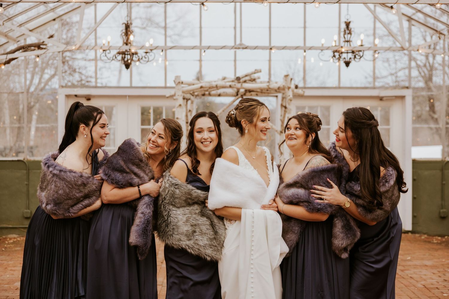 Bride with bridesmaids in navy gowns and fur wraps laughing outside winter greenhouse.