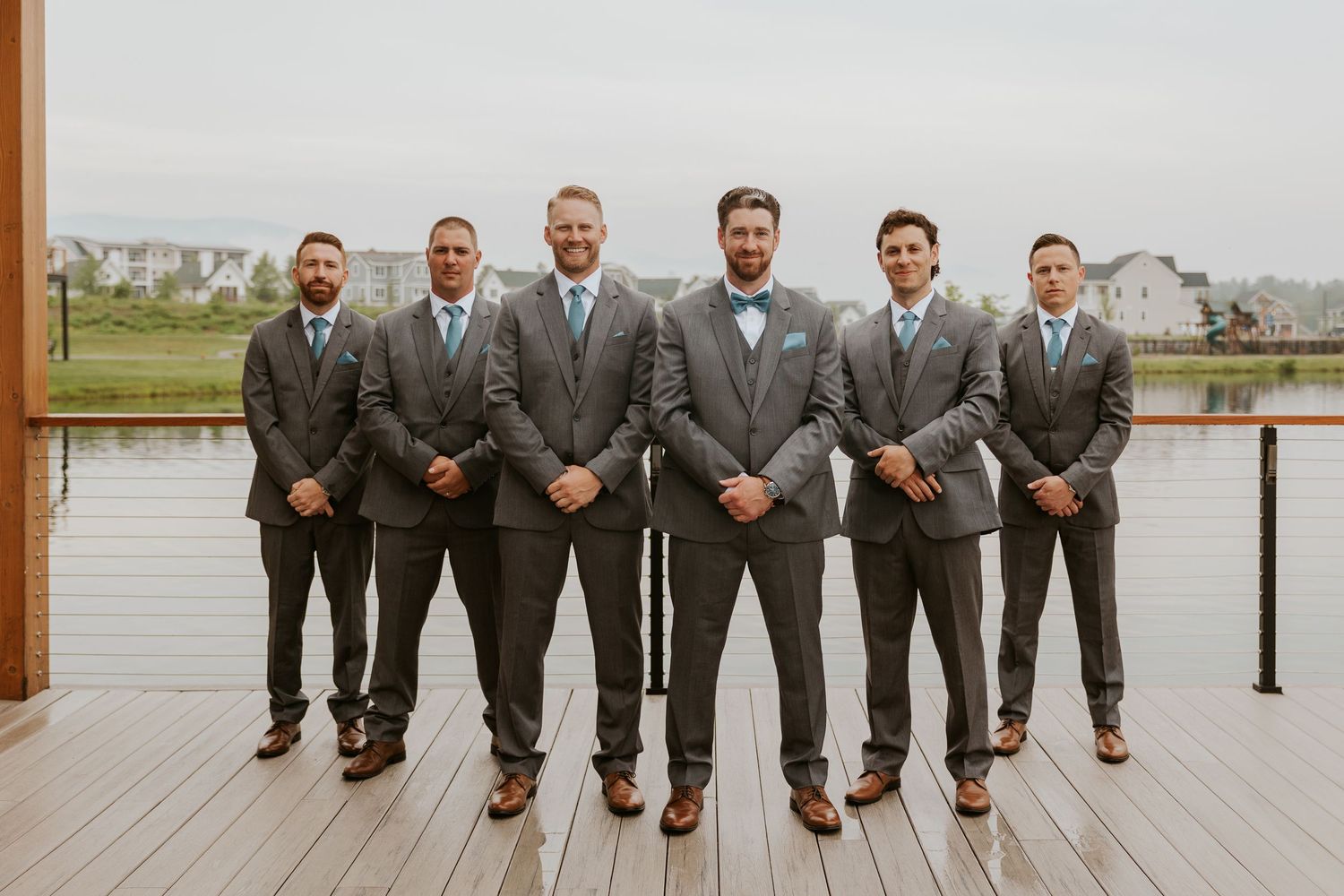 Groom and groomsmen in matching gray suits with blue bow ties posing on a wooden dock.