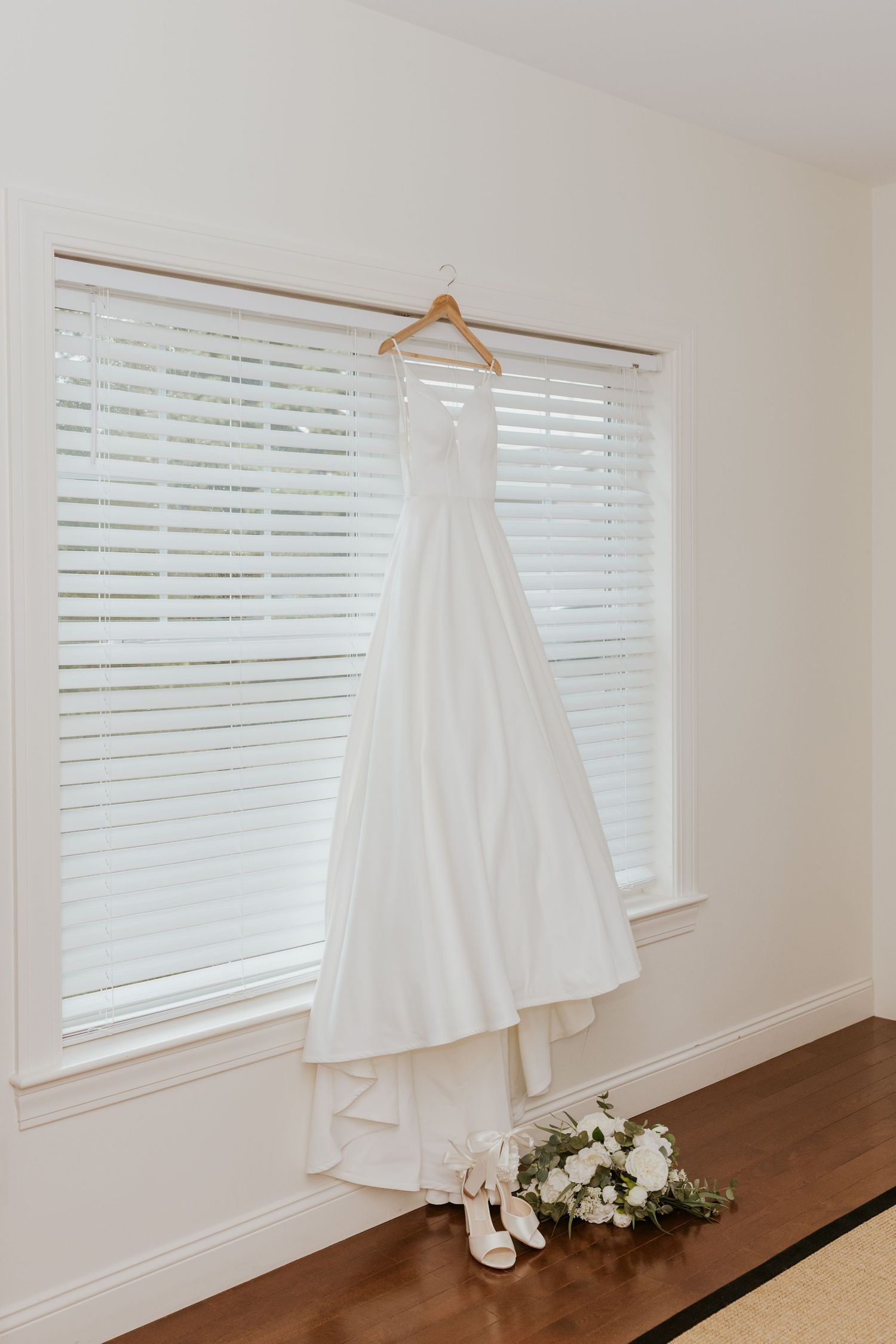 Elegant white wedding dress hanging by a window with bridal shoes and floral bouquet below.