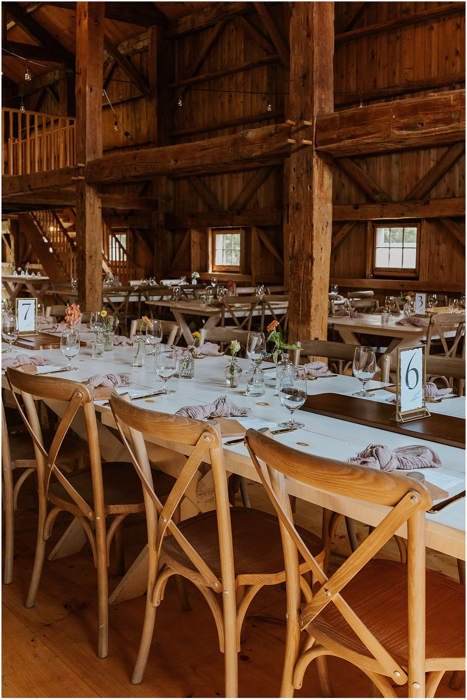Rustic barn wedding reception tables set with place settings and wooden cross-back chairs.