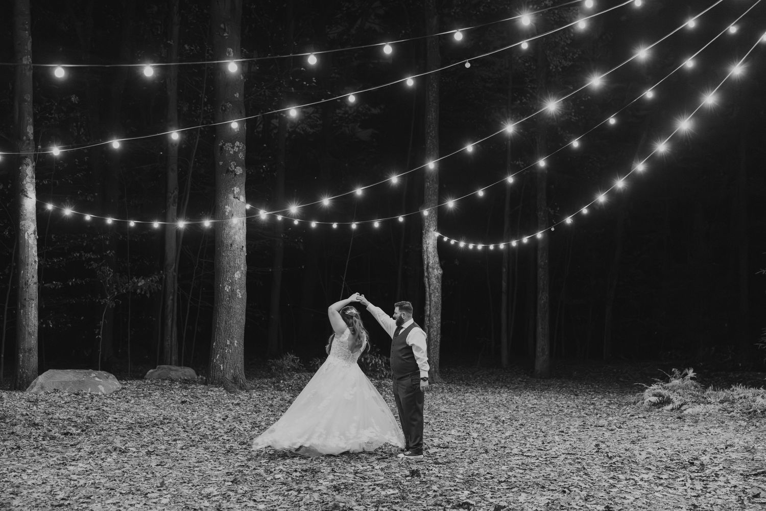 Bride and groom dance under twinkling string lights in forest at night in black and white photo.