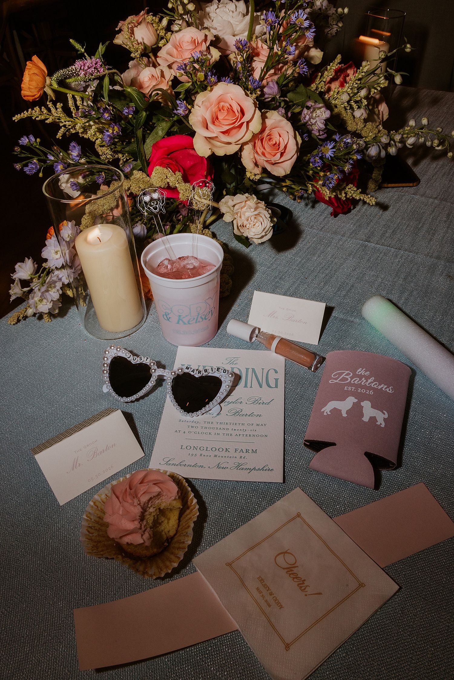 Wedding flat lay featuring floral bouquet, heart sunglasses, invitation suite, candle, and custom koozies on a table.