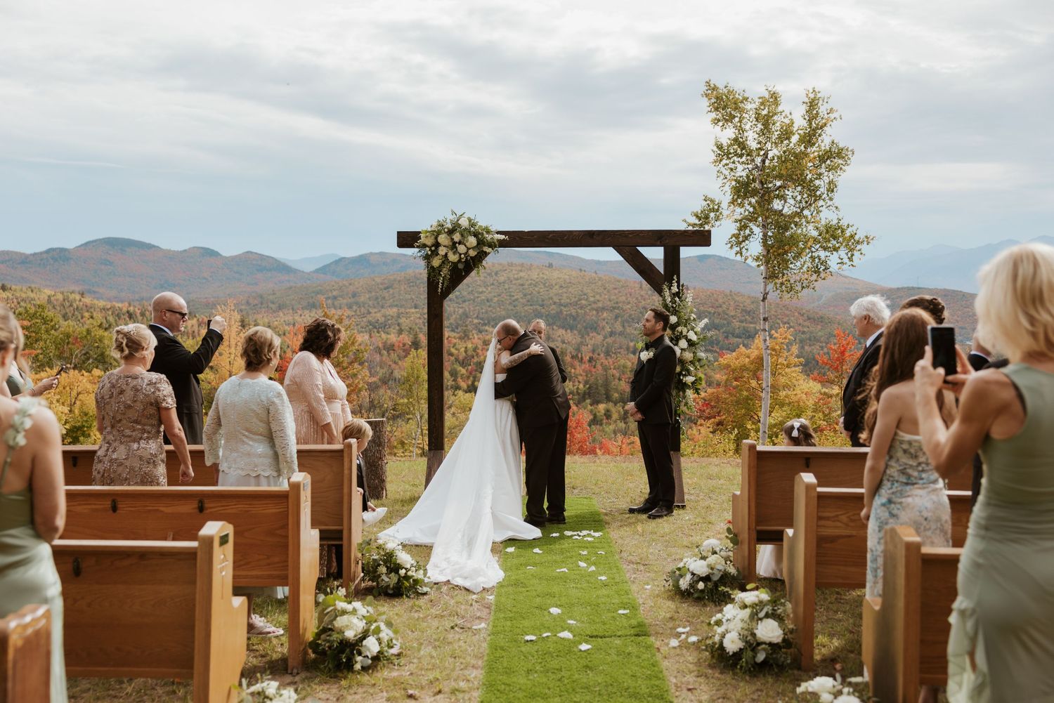 Outdoor fall wedding ceremony with couple kissing at altar, mountain backdrop and colorful foliage.