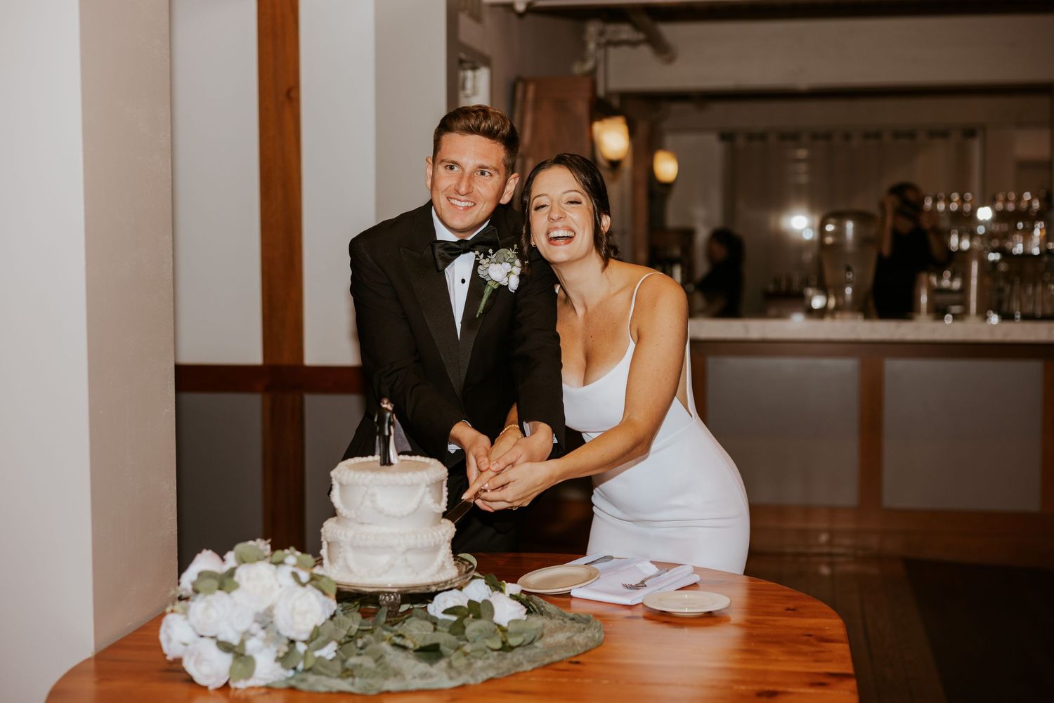 Smiling bride and groom cutting white wedding cake at reception.