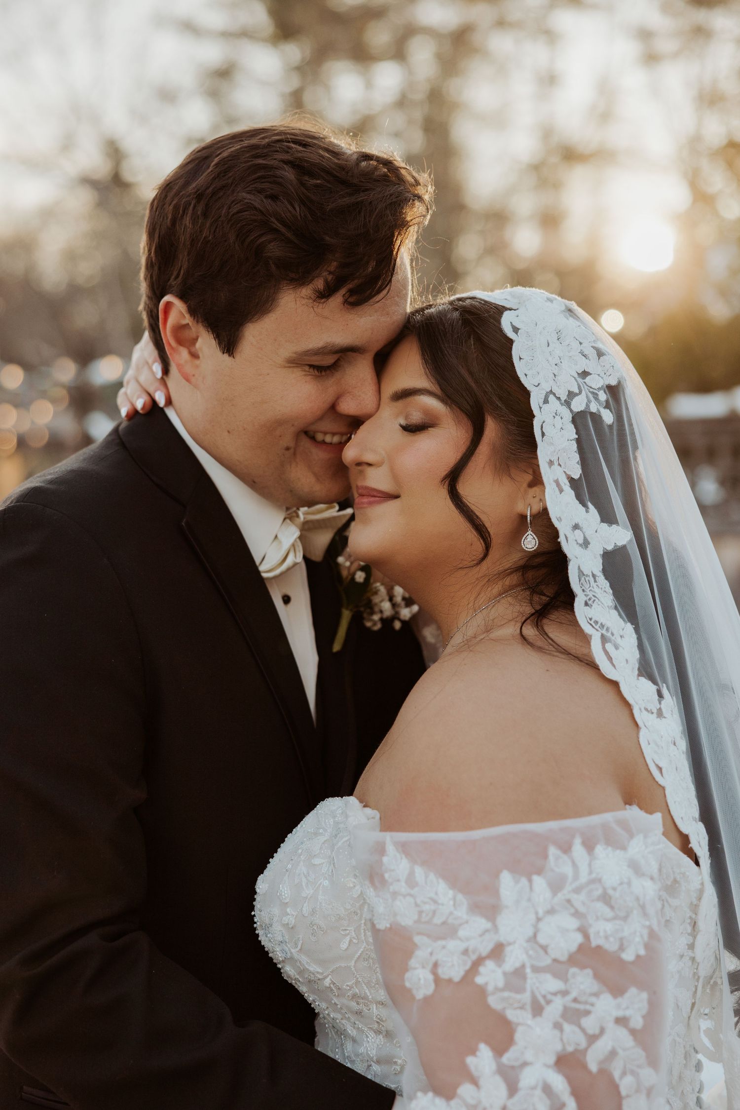 Bride and groom sharing a tender moment at sunset, bride wearing lace veil and off-shoulder gown.