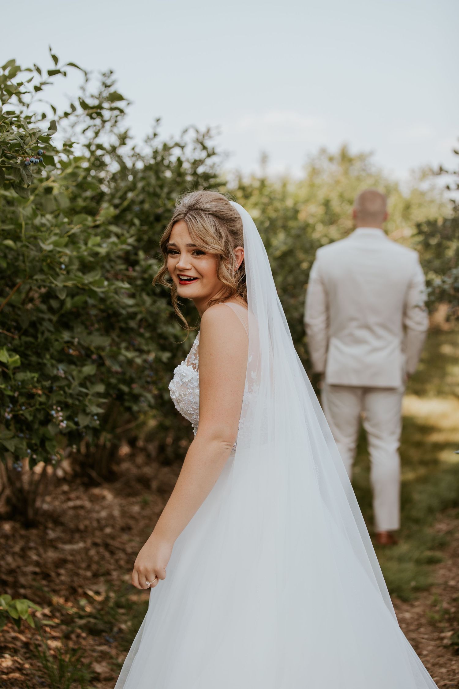 Bride in a white lace gown and veil smiling back at camera while groom stands ahead in an orchard.