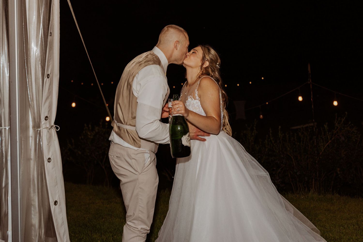 Bride and groom sharing a kiss while holding a champagne bottle at outdoor night wedding.