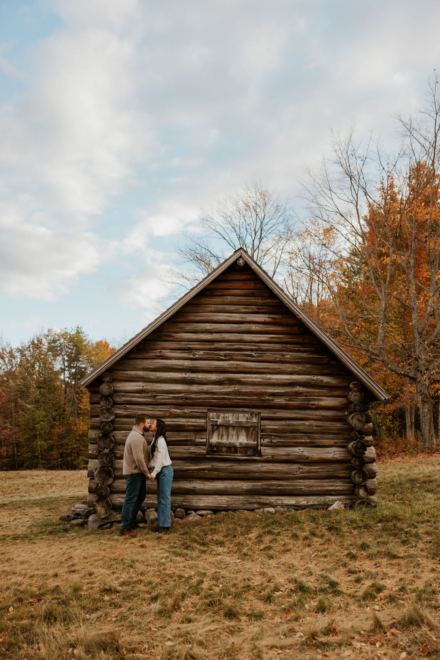 Couple leaning together beside a rustic log cabin in autumn landscape.