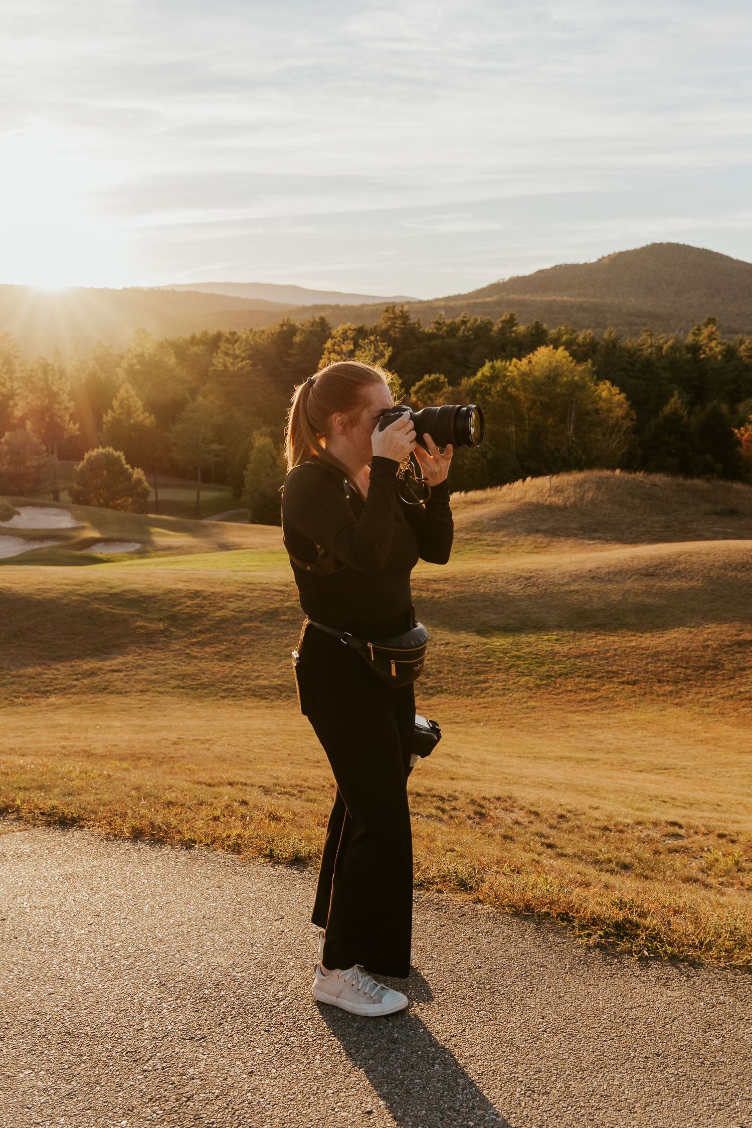 Female photographer captures shots at golden hour on a scenic golf course with mountains and autumn foliage behind her.