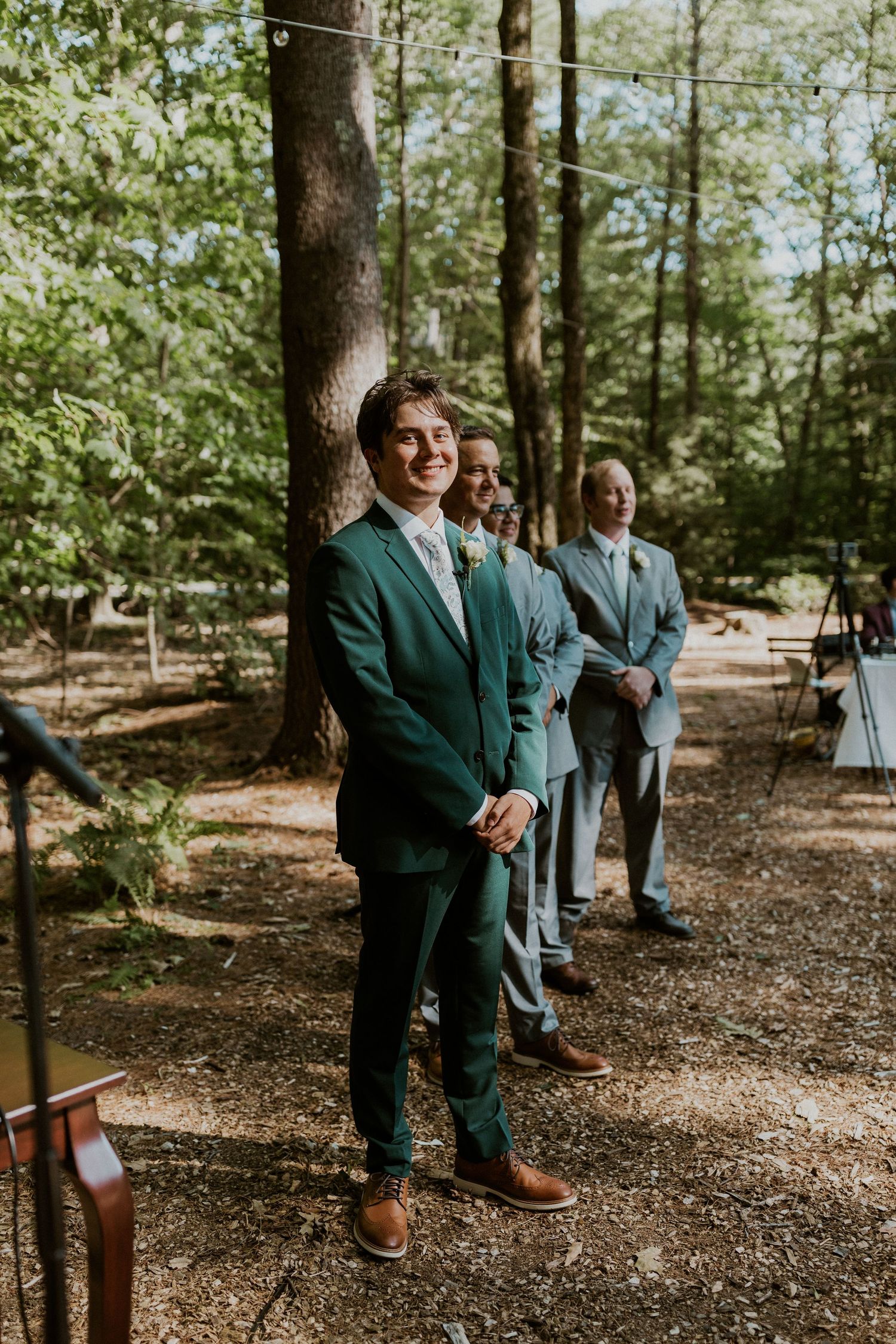 Wedding party members in green and grey suits stand together in a forest setting.