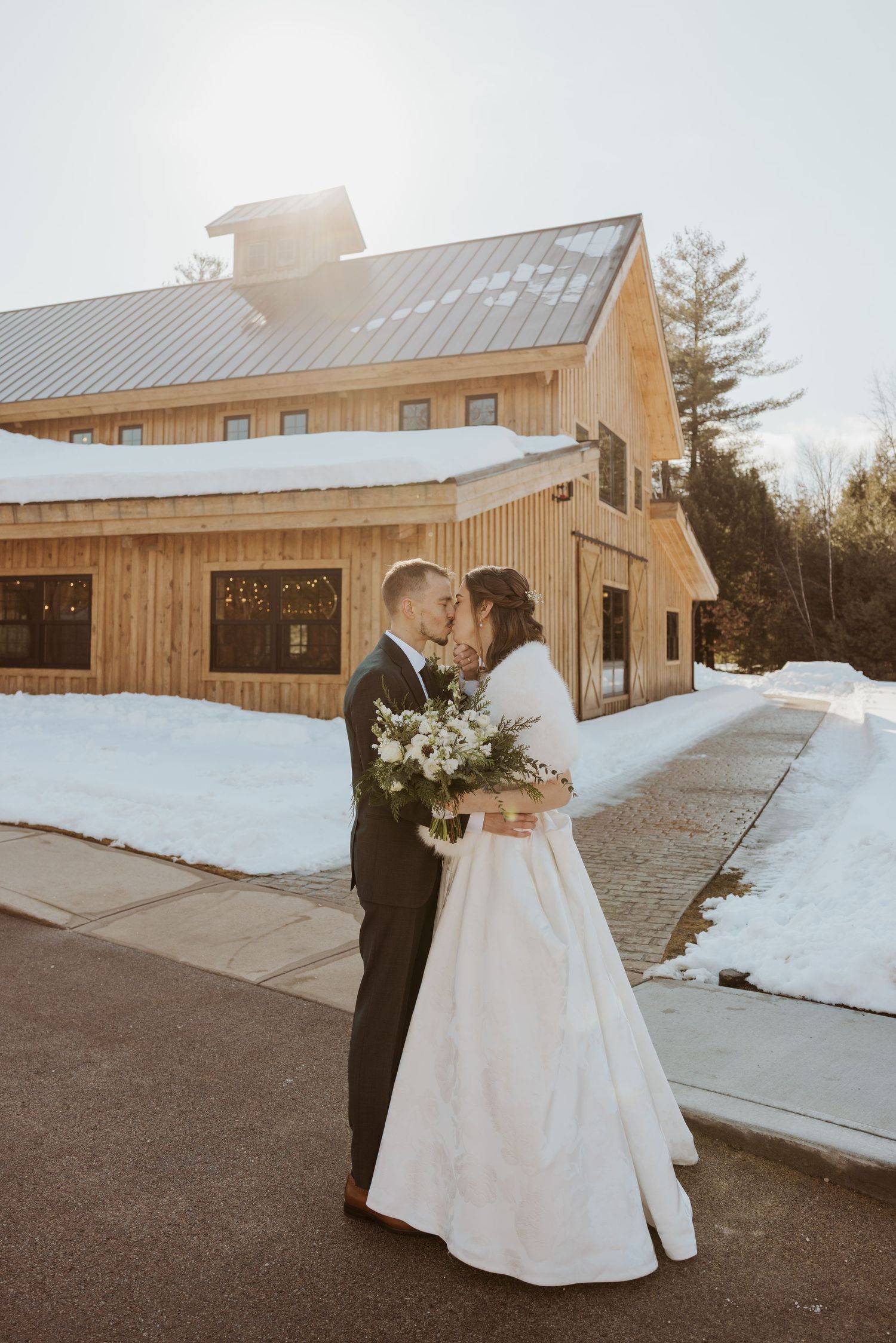 A couple in wedding attire embraces outside a rustic wooden barn venue surrounded by snowy winter landscape.