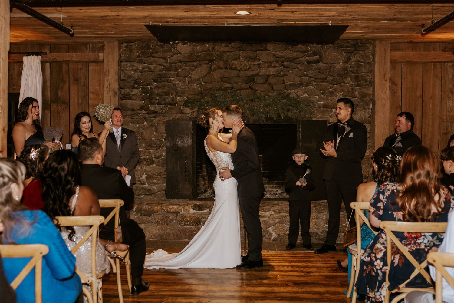 Bride and groom share first kiss at indoor wedding ceremony beside stone fireplace.