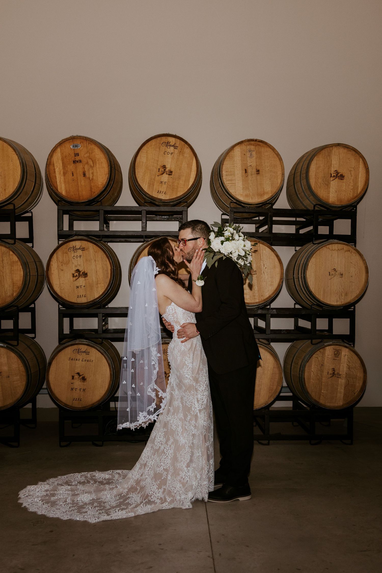 A romantic wedding photo in a winery with wooden barrels stacked on racks in the background.