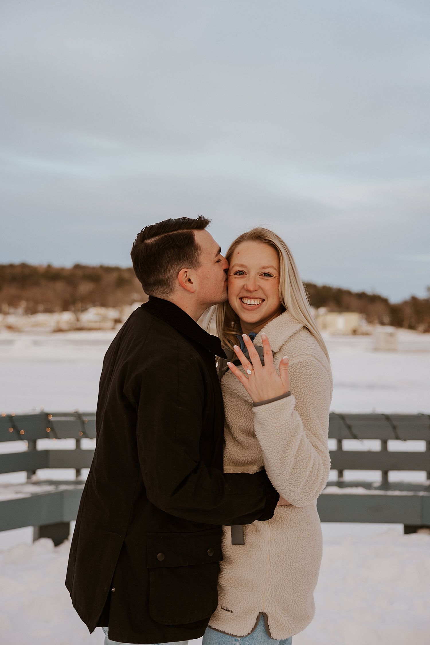 Couple celebrating engagement in snowy winter setting, woman showing ring while man kisses her cheek.