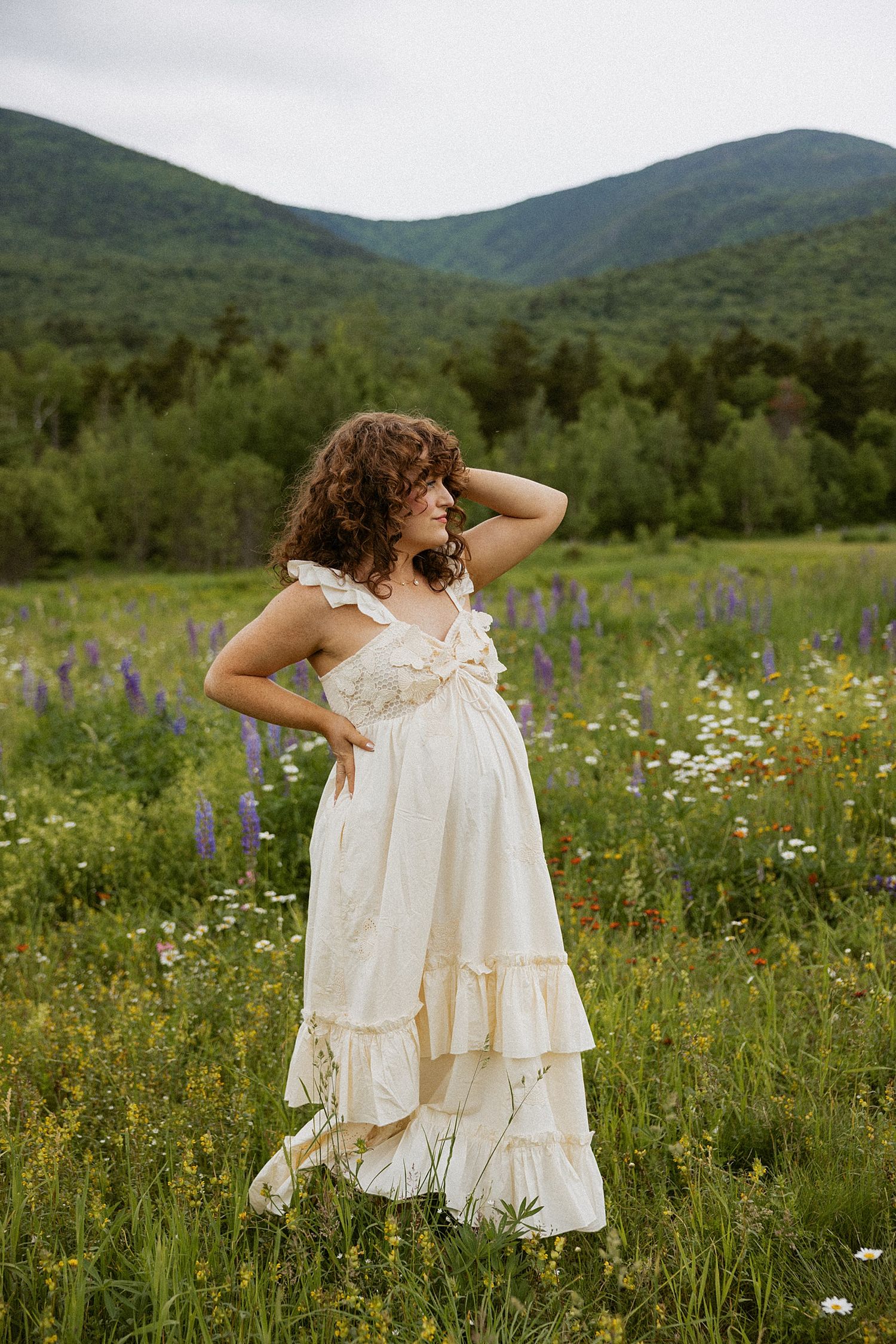 Pregnant woman in white ruffled dress standing in wildflower field in NH taking motherhood photos