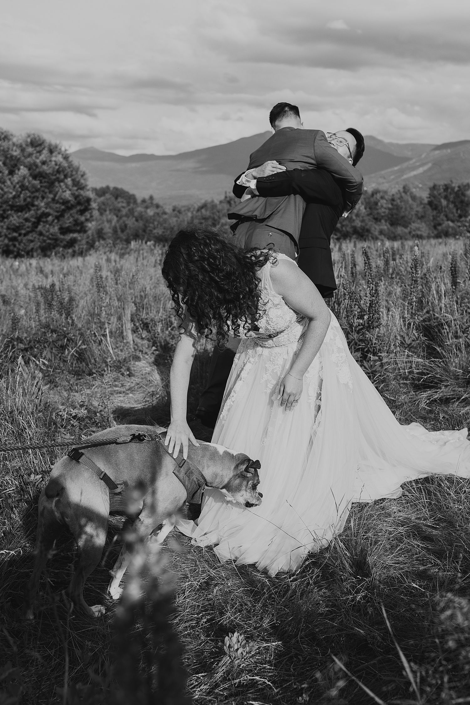 Black and white photo of bride petting dog while groom embraces her in scenic mountain meadow landscape.