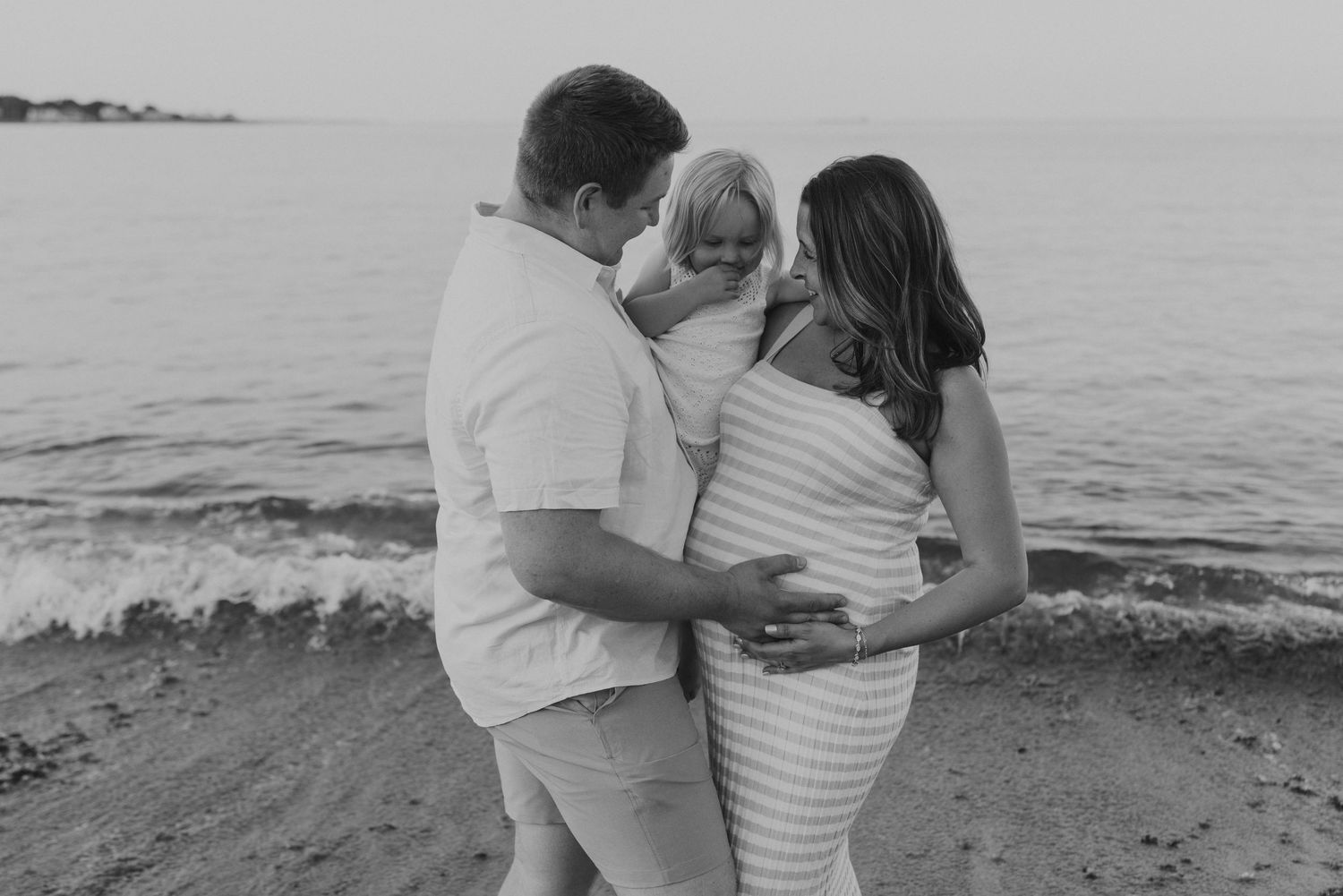 Black and white photo of pregnant couple holding toddler on beach with ocean waves behind them.