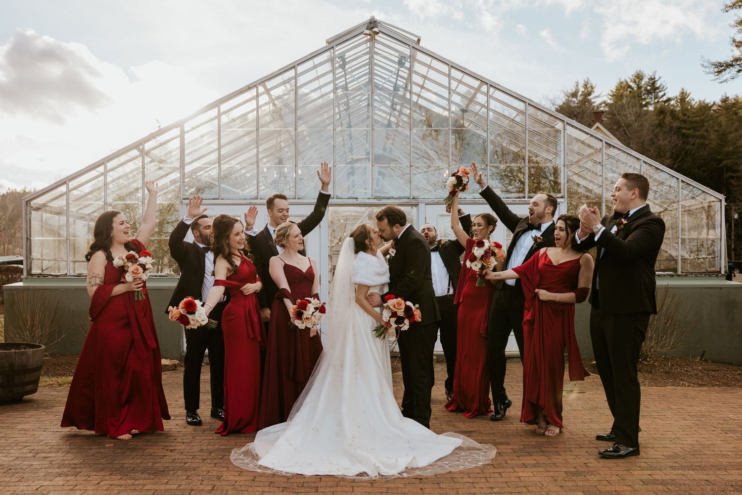 Wedding party in black tuxedos and red dresses celebrate outside a glass greenhouse venue.