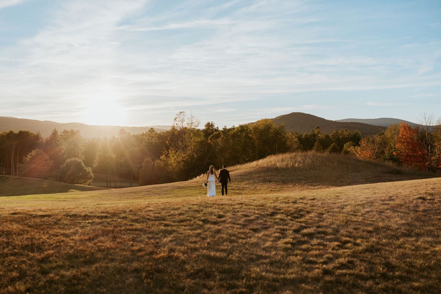 Bride and groom walk through vast golden meadow with stunning mountain sunset landscape backdrop.