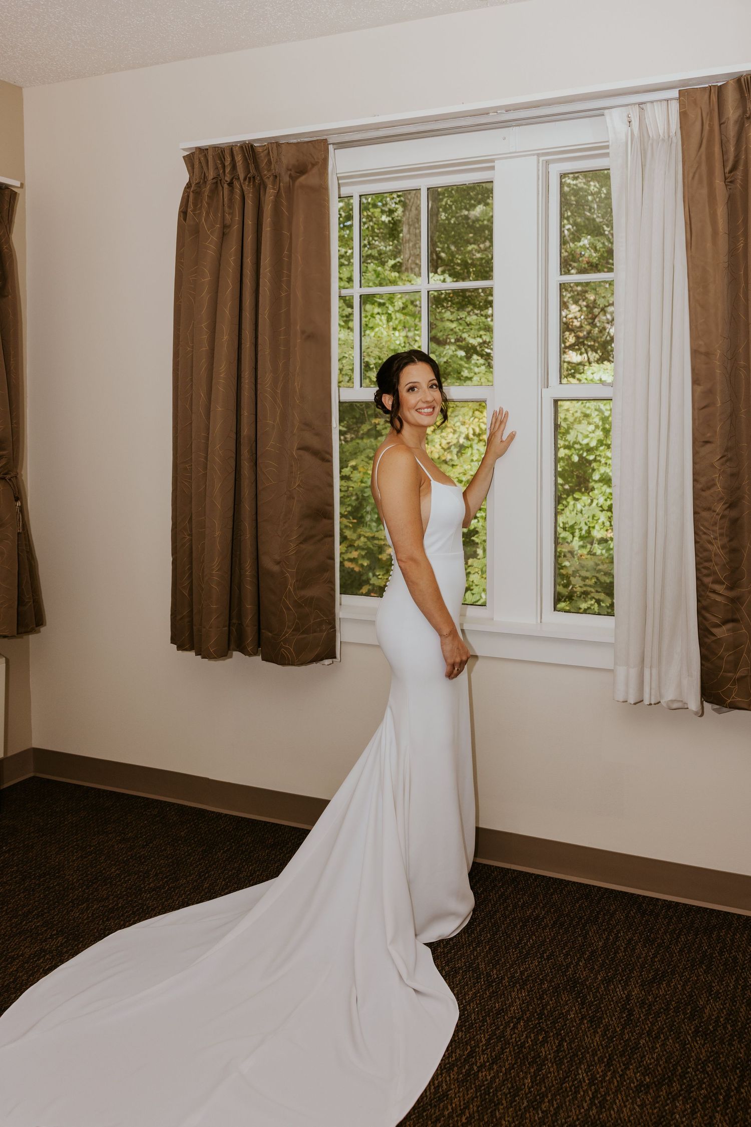 Bride in elegant white fitted gown with long train standing by window in hotel room on wedding day.