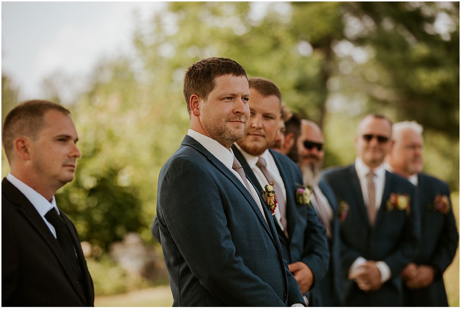 Groomsmen in dark suits standing together outdoors during wedding ceremony.