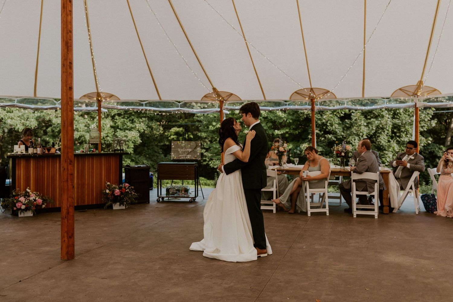 Wedding reception setup under a white tent with string lights and greenery decorations.
