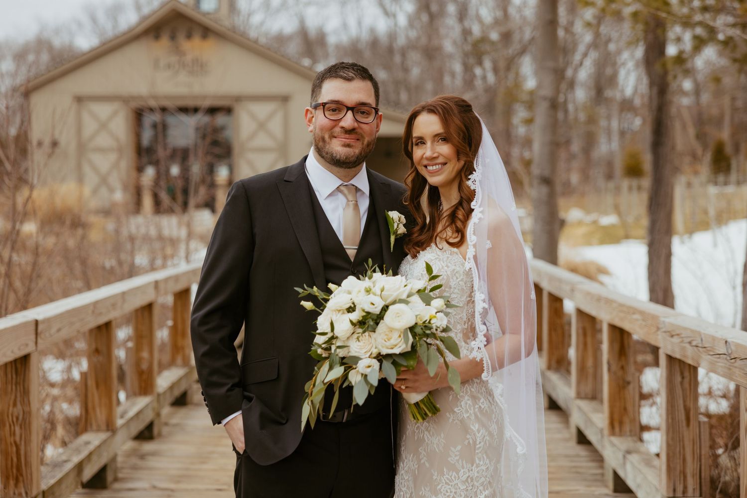 Winter wedding couple poses on a rustic wooden bridge surrounded by snow-covered trees and white floral arrangements.