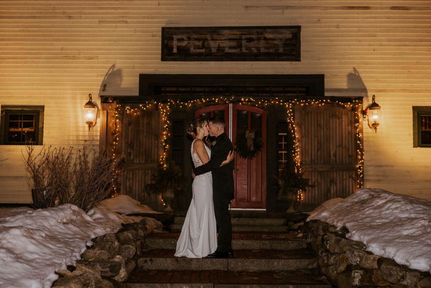 Bride and groom sharing a kiss outside a snow-covered rustic venue at night.