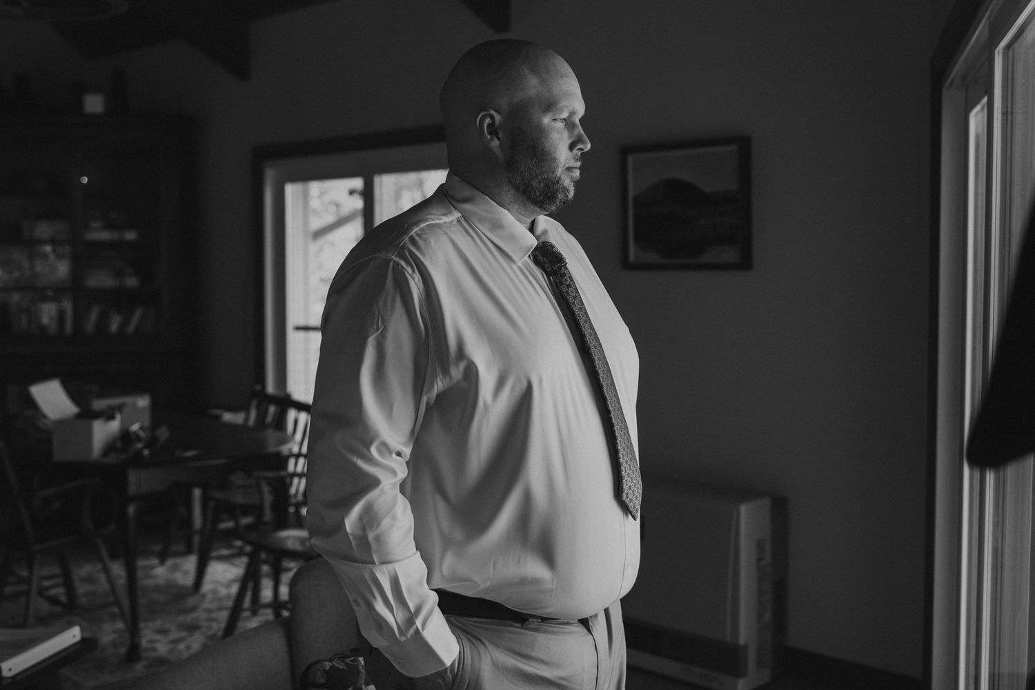 A black and white photograph shows a person in a light colored dress shirt and tie standing in a dimly lit room.