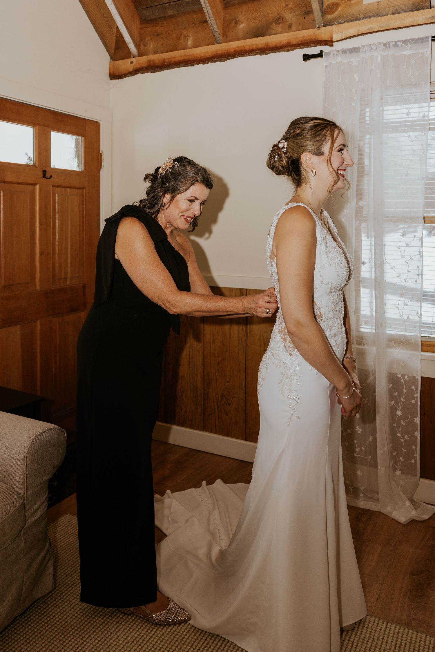Mother helping bride zip up elegant white lace wedding gown in rustic cabin room.