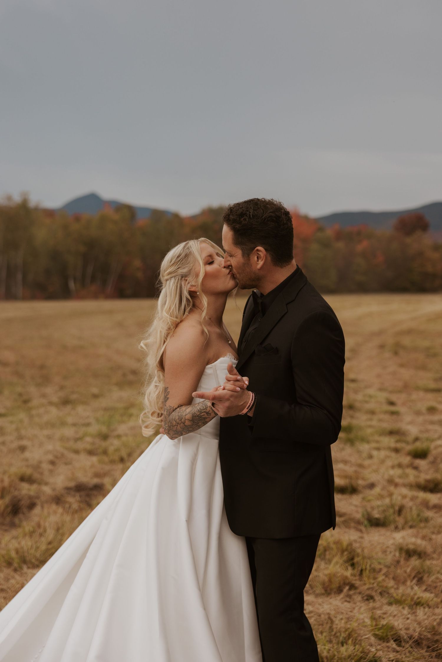 Bride and groom sharing first kiss in autumn field with colorful fall foliage backdrop.